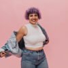 Fashionable lady in stylish tight jeans and short white top looks at camera on pink background. Funny girl with violet hair takes off her jacket. Para look de oficina o salidas