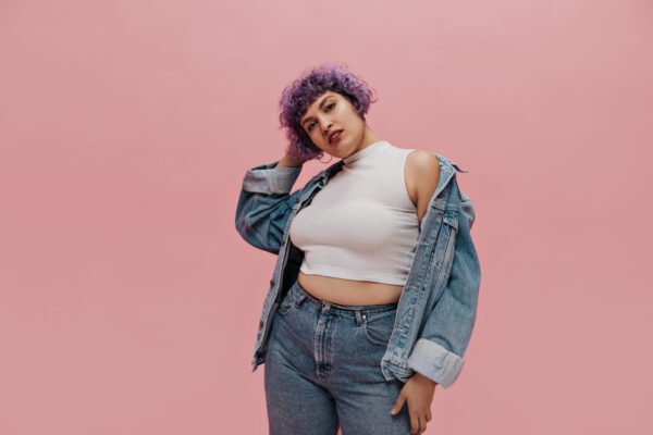 Cool woman with short multi-colored hair in round earrings, white T-shirt, large denim jacket and in light pants looks at camera on pink background. Para look de oficina o salidas