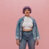 Smiling adult woman with short curly purple hair in white top, in denim jacket and tight jeans posing on isolated background. Para look de oficina o salidas