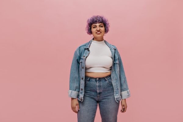Smiling adult woman with short curly purple hair in white top, in denim jacket and tight jeans posing on isolated background. Para look de oficina o salidas
